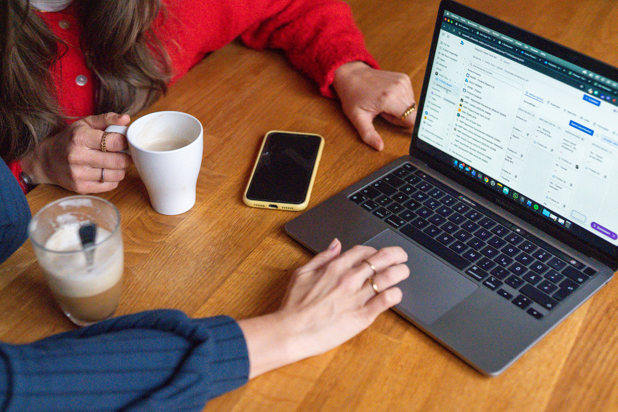 Person hält eine Tasse Kaffee und hat ein Smartphone auf dem Tisch neben einem Laptop